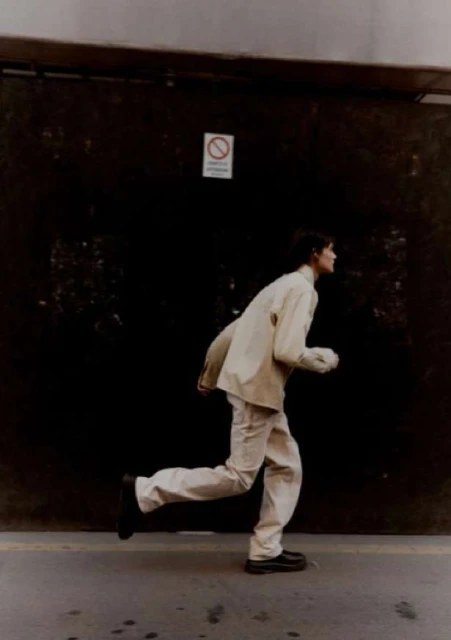 Full shot of a slender, pale-skinned individual in a casual tan jacket and pants running on a gray concrete sidewalk against a dark wall.