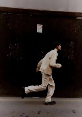 Full shot of a slender, pale-skinned individual in a casual tan jacket and pants running on a gray concrete sidewalk against a dark wall.