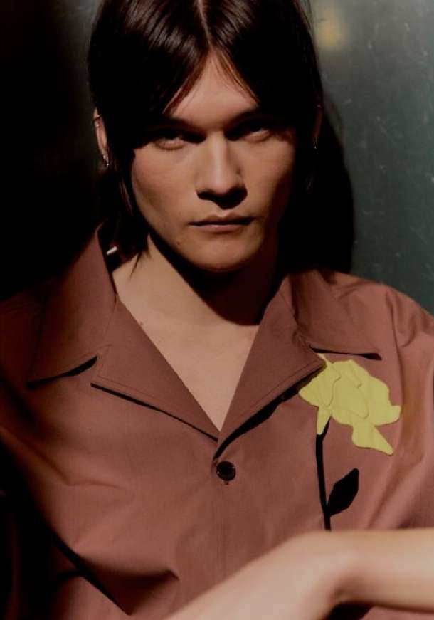 Portrait of a young adult with brown hair wearing a mauve button-down shirt with a yellow flower applique, indoor lighting.