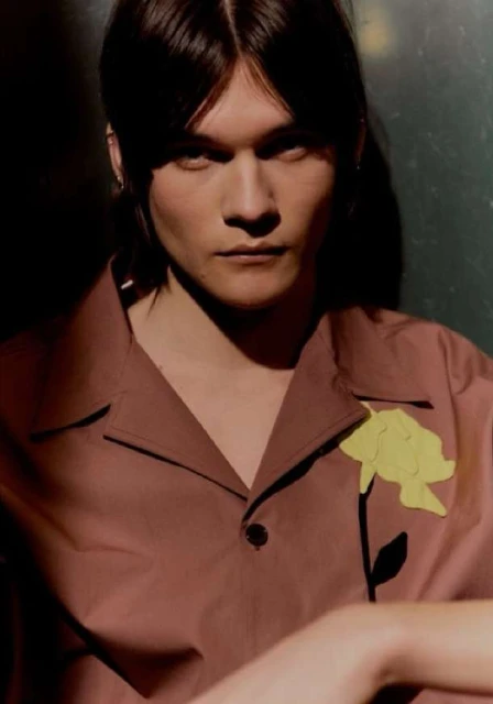 Portrait of a young adult with brown hair wearing a mauve button-down shirt with a yellow flower applique, indoor lighting.