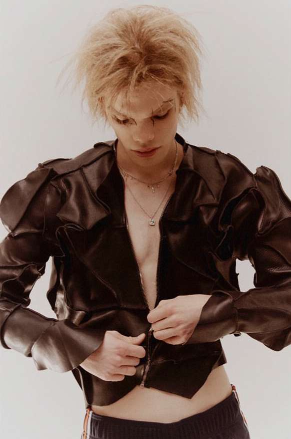 Close-up portrait of a young adult with fair skin and blonde hair wearing a black leather jacket and jewelry against a white background.