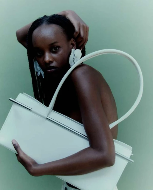 A fashionable dark-skinned woman poses with a white handbag in a studio setting, highlighted by her flower earrings and braided hair.