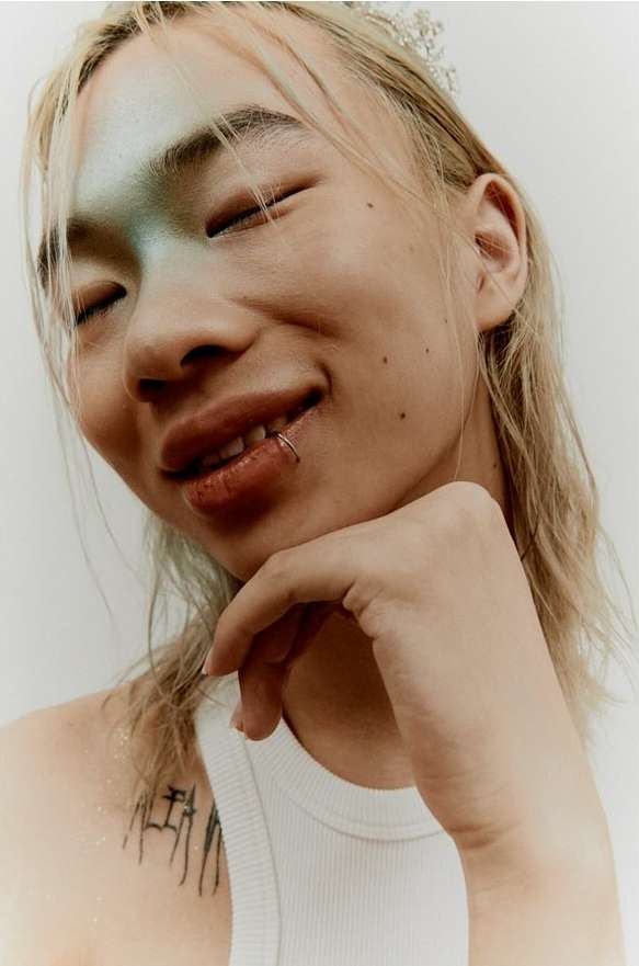 Close-up portrait of a smiling adult with lip ring, pale skin, blonde hair, and a white tank top in a photo studio setting.