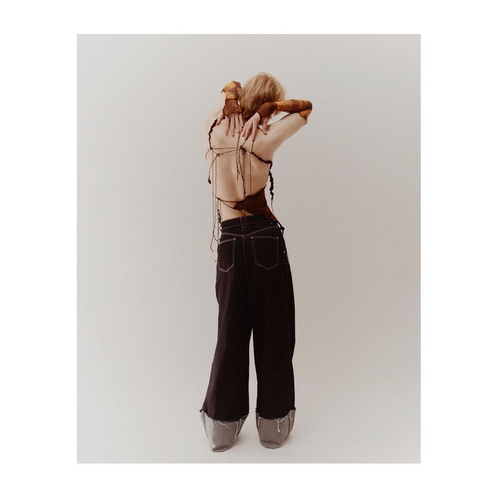 A woman stands with her back to the camera, wearing an open-back top and dark denim pants against a neutral studio backdrop.