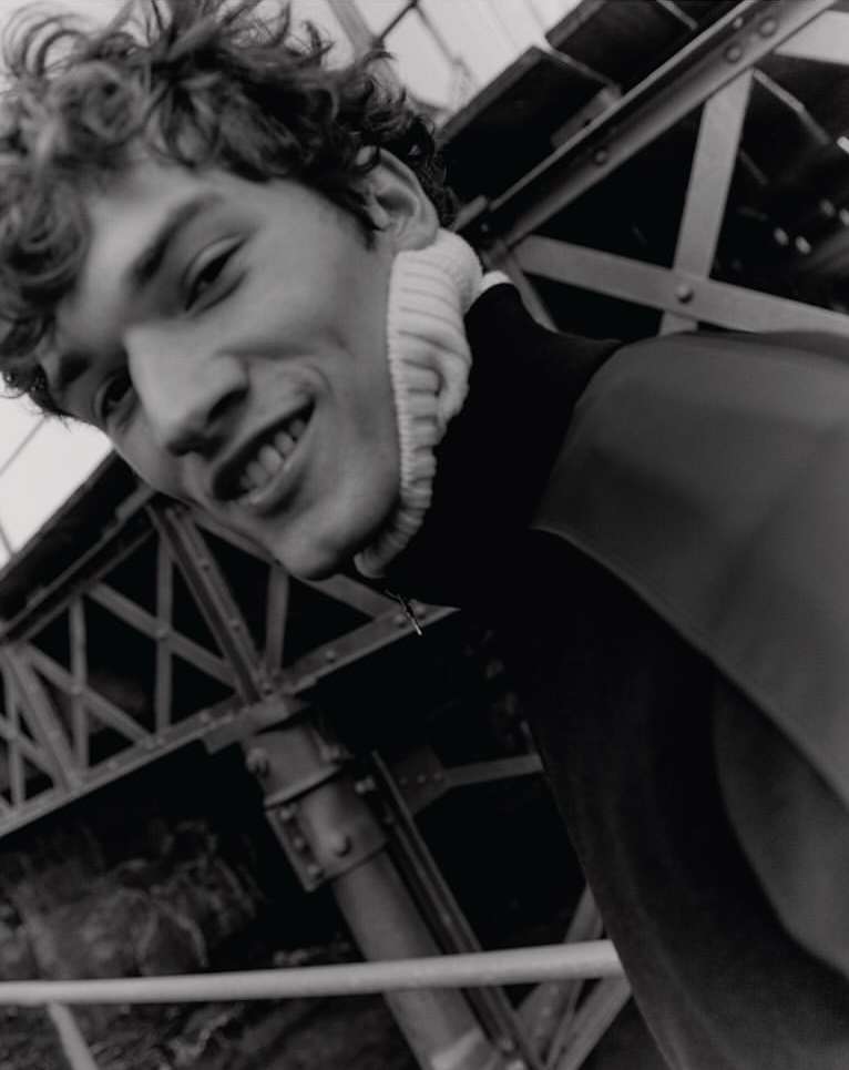 Black and white portrait of a smiling young adult male with curly hair, wearing a high-neck sweater, with an industrial background.