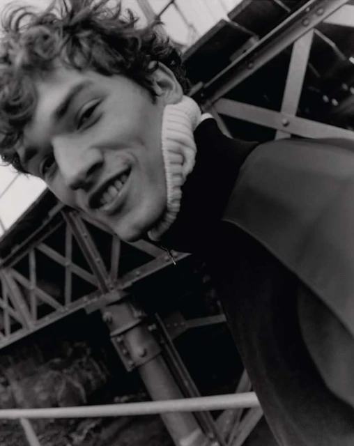 Black and white portrait of a smiling young adult male with curly hair, wearing a high-neck sweater, with an industrial background.