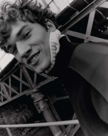 Black and white portrait of a smiling young adult male with curly hair, wearing a high-neck sweater, with an industrial background.