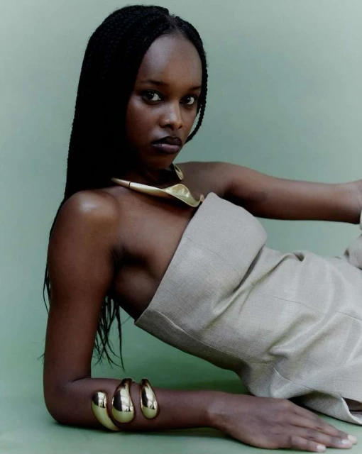 A woman with dark skin and long braided hair poses in a strapless dress with gold jewellery against a light green background in a studio shot.