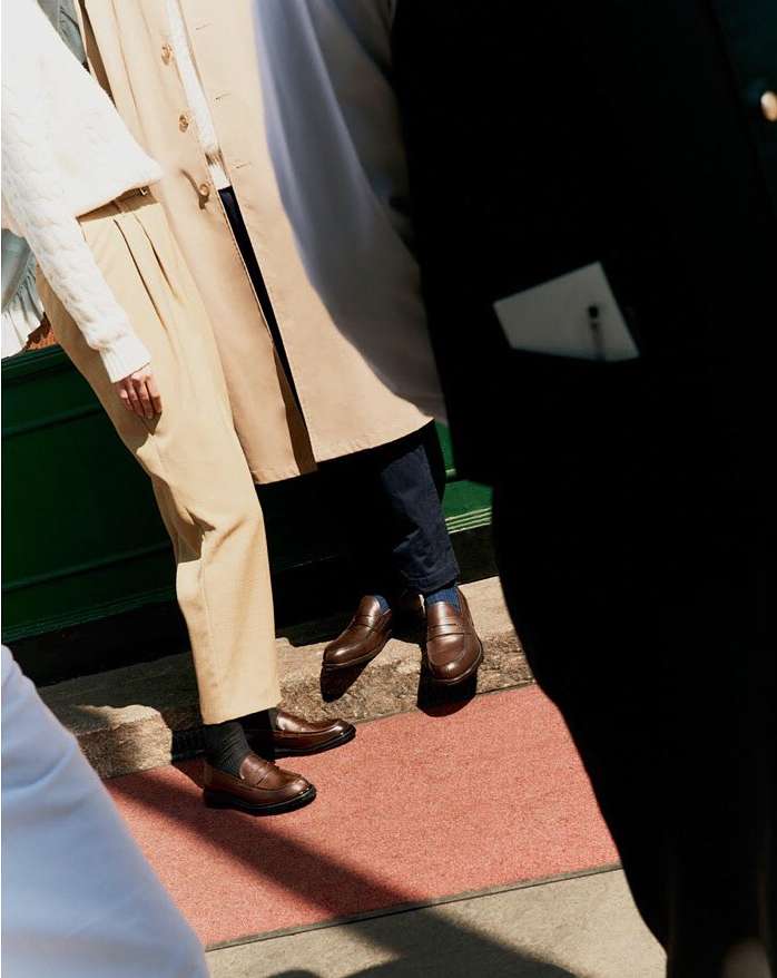 A group of people wearing smart casual outfits, including dress shoes and trousers, stand on a red rug, in a brightly lit outdoor setting.