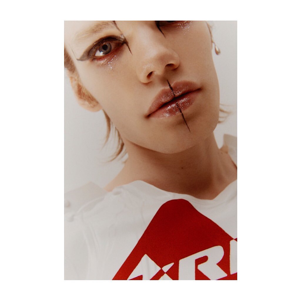Close-up portrait of a fair-skinned adult woman with graphic black eyeliner and glossy lips, wearing a white t-shirt indoors.