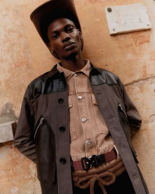 Adult male in a brown cowboy hat, brown shirt, black jacket, and brown pants leaning against a peach colored wall for a portrait.