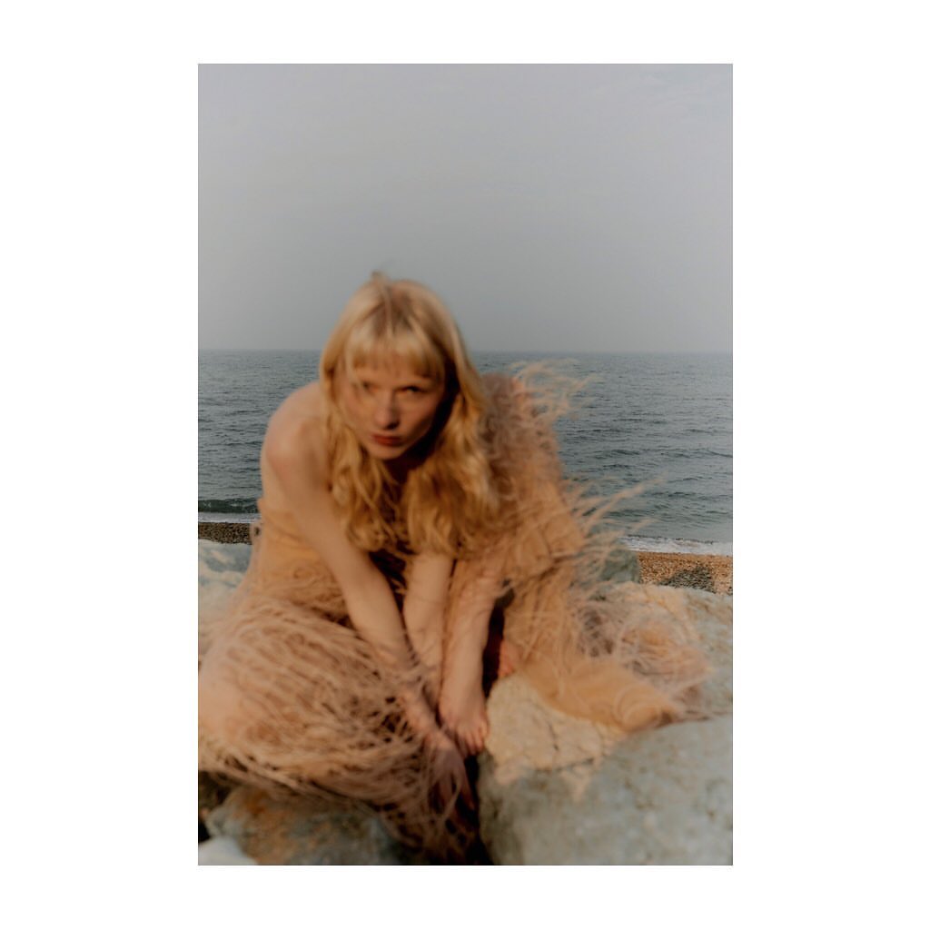 Blonde woman in flowing peach gown poses on rocks near the ocean, staring intently into the camera, with a moody, atmospheric sky.
