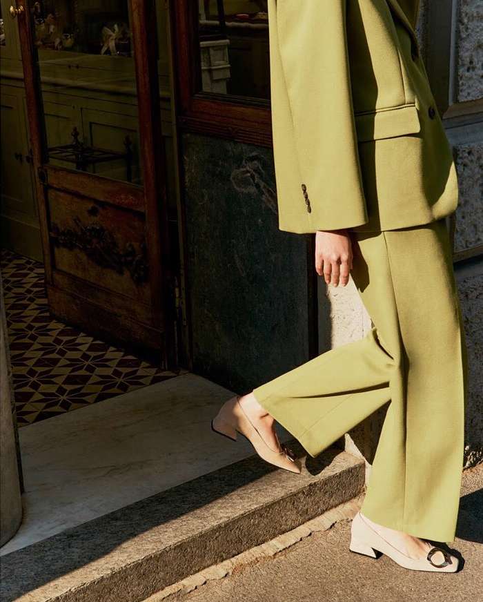 Woman in a green suit walks out of an old doorway, showcasing beige block-heeled shoes and sunlight casting shadows on the ground.