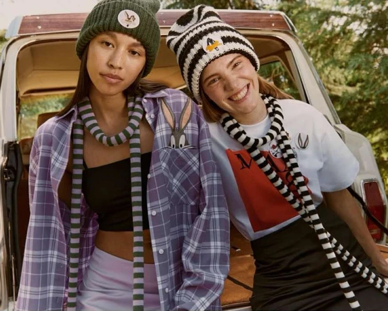 Two smiling women in hats pose outdoors near a car, showcasing cartoon character apparel in a friendly, casual setting. Fashionable and fun.