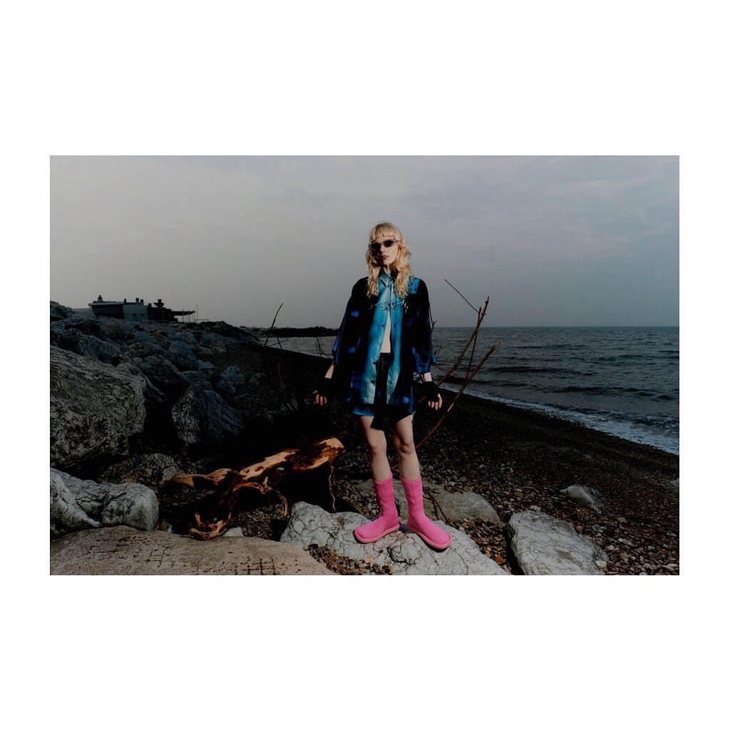 Fashion shot of a blonde adult woman in an open patterned coat and pink boots, standing on a rocky beach on a cloudy day.