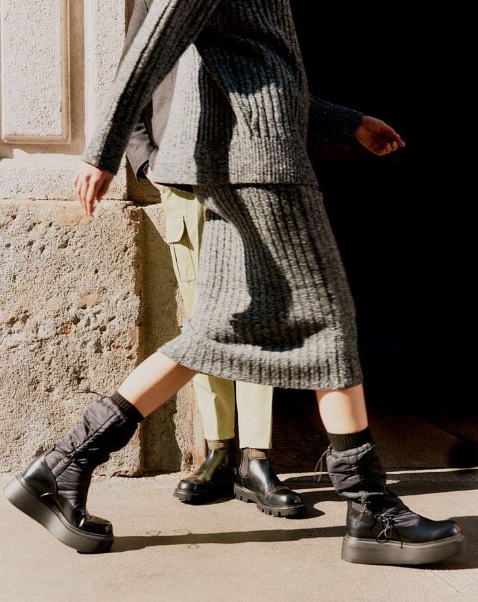 Two people wearing neutral-toned knitwear and black platform boots walk past a concrete wall in an outdoor urban setting.