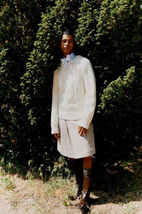 Man in white cable knit sweater and skirt stands in a forest setting with tall trees and dappled sunlight, modeling fashion.