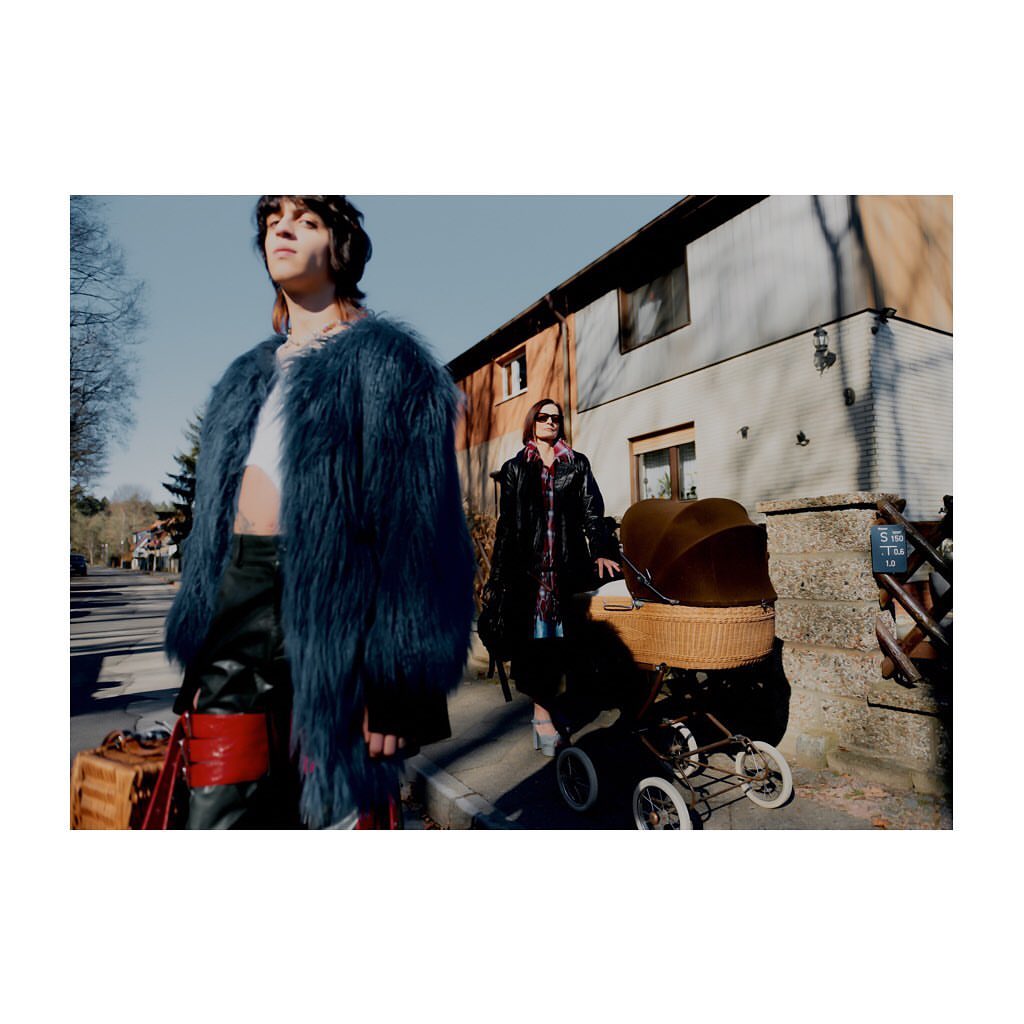 Two people walking down a sidewalk, one pushing a vintage baby carriage, while the other wears a blue fur coat and carries luggage.