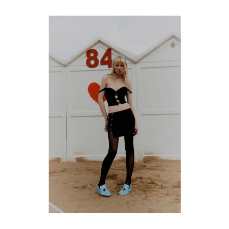 Blonde model in a black off-the-shoulder crop top and skirt with blue loafers standing on sand before beach cabins.
