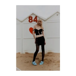 Blonde model in a black off-the-shoulder crop top and skirt with blue loafers standing on sand before beach cabins.