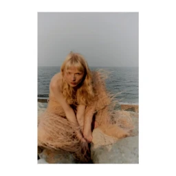 Blonde woman in a textured sheer beige dress crouching on rocky coastal shore against an ocean background