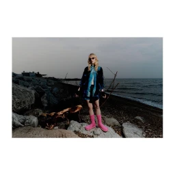 Model wearing dark blue patterned jacket and pink boots standing on a rocky shoreline by the ocean