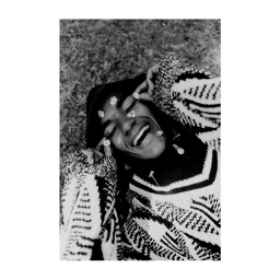 Black and white portrait of a smiling person wearing a patterned sweater with small flowers placed on their face, lying on grass.