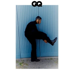 Man wearing all black outfit and black boots stepping up against a vertical blue paneled wall photographed in Milan by Pietro Groff.