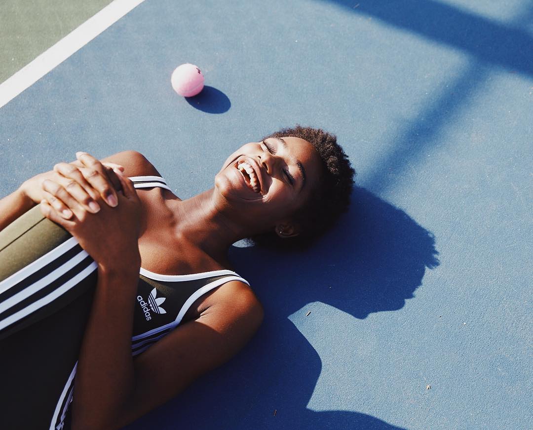 Smiling woman in athletic wear lying on a blue tennis court with a tennis ball, expressing joy and health in bright sunlight.