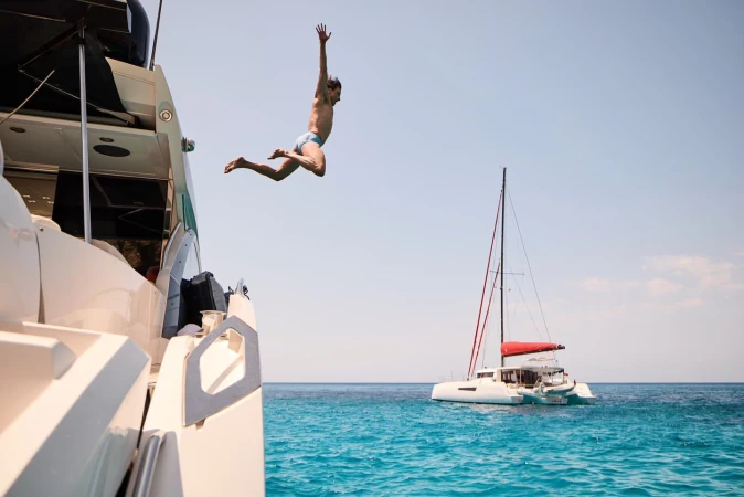 An adult man in mid-air jumping off a large white yacht into turquoise ocean water, with a white catamaran sailboat in the background.