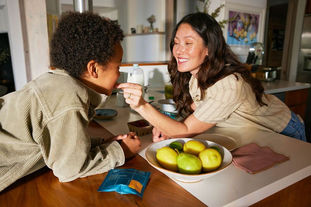 Woman feeding a berry to a child in a bright kitchen with lemons, limes, and pears in a bowl on the counter. Healthy eating.