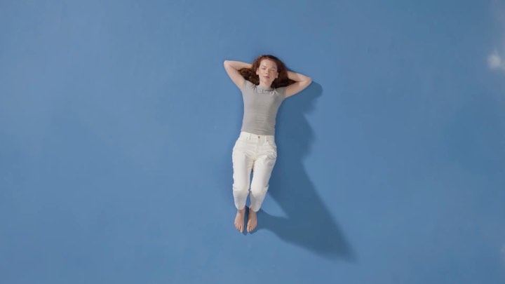 Overhead shot of a redhead woman in white jeans and a gray shirt laying on a vibrant blue surface with hands behind her head.