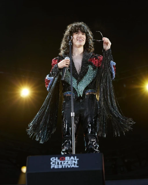 Performer singing at Global Citizen Festival: A singer with long curly hair holds a microphone on stage with stage lighting.