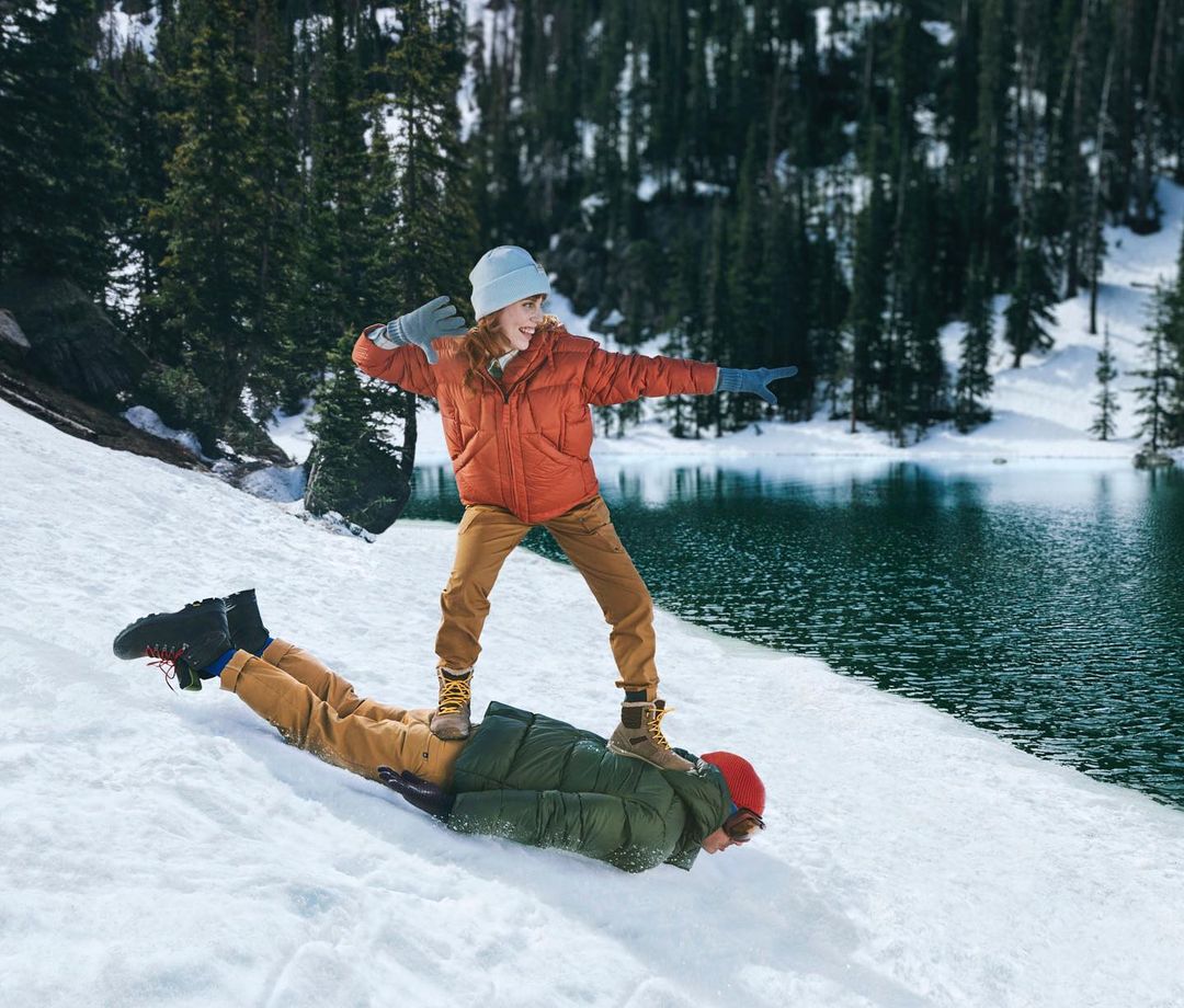 A young person stands on the back of someone lying in snow near a lake, with evergreen trees in the background on a sunny day.