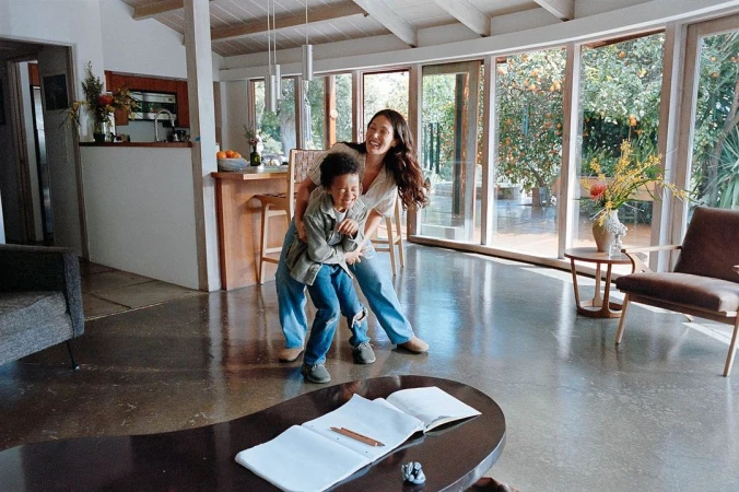 Mother and child laughing and embracing in a modern living room with large windows overlooking an outdoor garden with fruit trees.