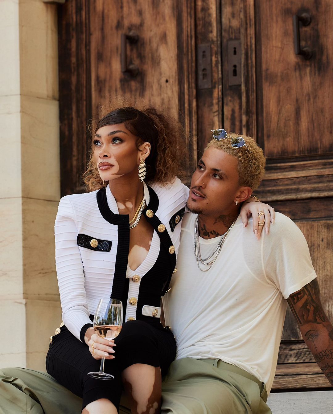Model couple in elegant outdoor setting, woman in black and white blazer holding wine, man in white t-shirt and tattoos.