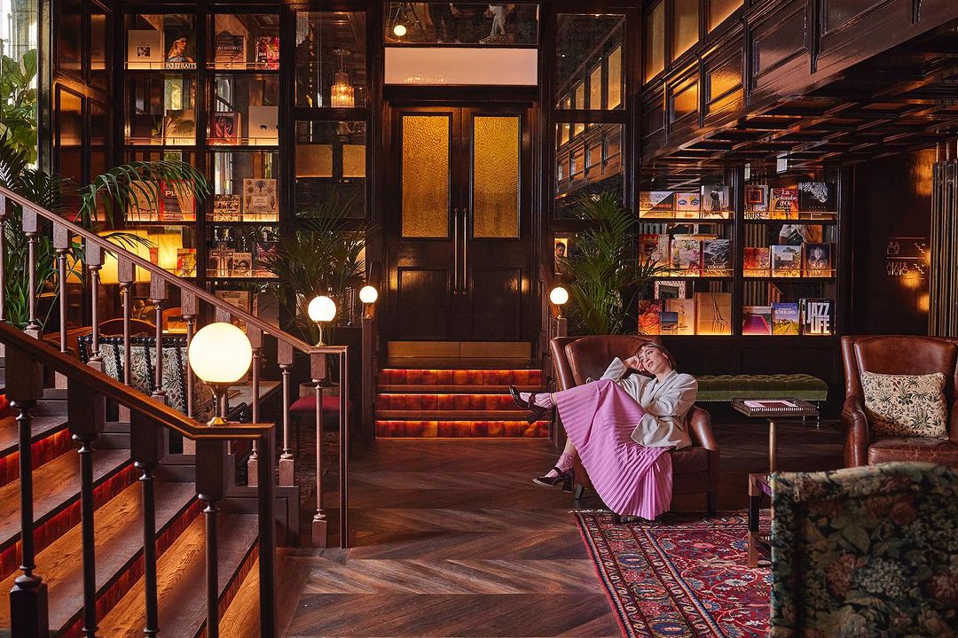 Woman in armchair in library-style lounge with bookshelves, herringbone floors, and warm lighting.