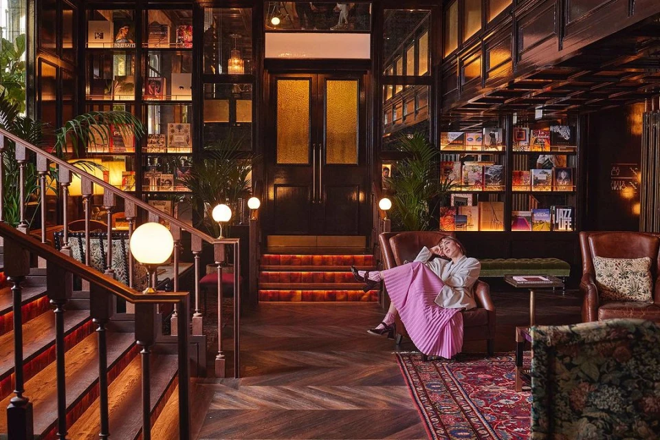 Woman in armchair in library-style lounge with bookshelves, herringbone floors, and warm lighting.