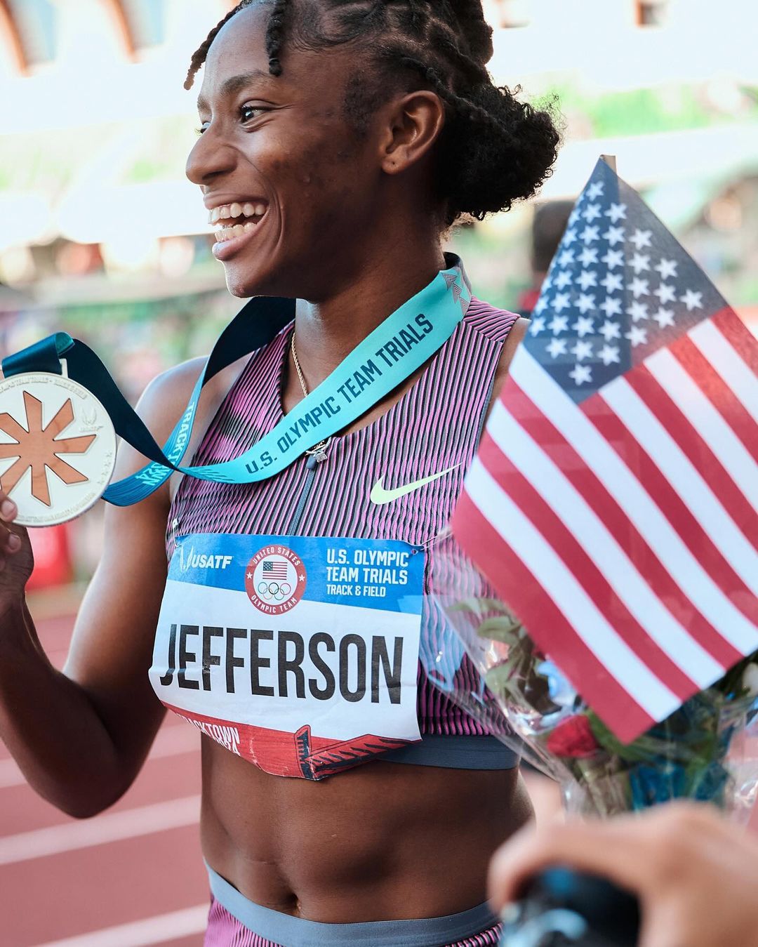 Athlete celebrates U.S. Olympic Team Trials victory with American flag, wearing medal, smiling outdoors in stadium.