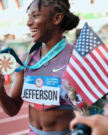 Athlete celebrates U.S. Olympic Team Trials victory with American flag, wearing medal, smiling outdoors in stadium.