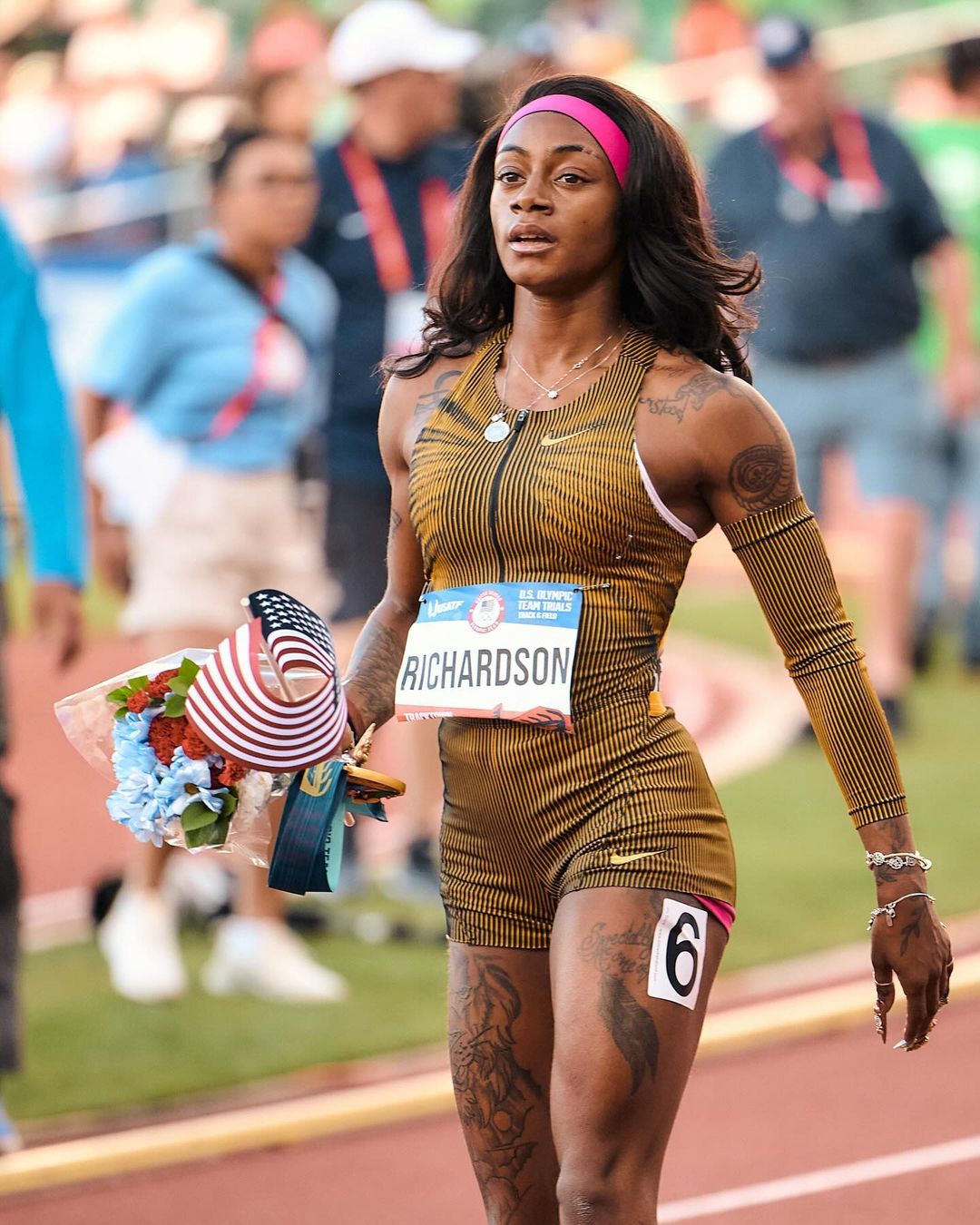 Sha'Carri Richardson, an adult female athlete with tattoos, stands on a track with a bouquet of flowers and an American flag.