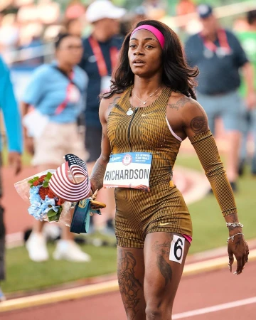 Sha'Carri Richardson, an adult female athlete with tattoos, stands on a track with a bouquet of flowers and an American flag.