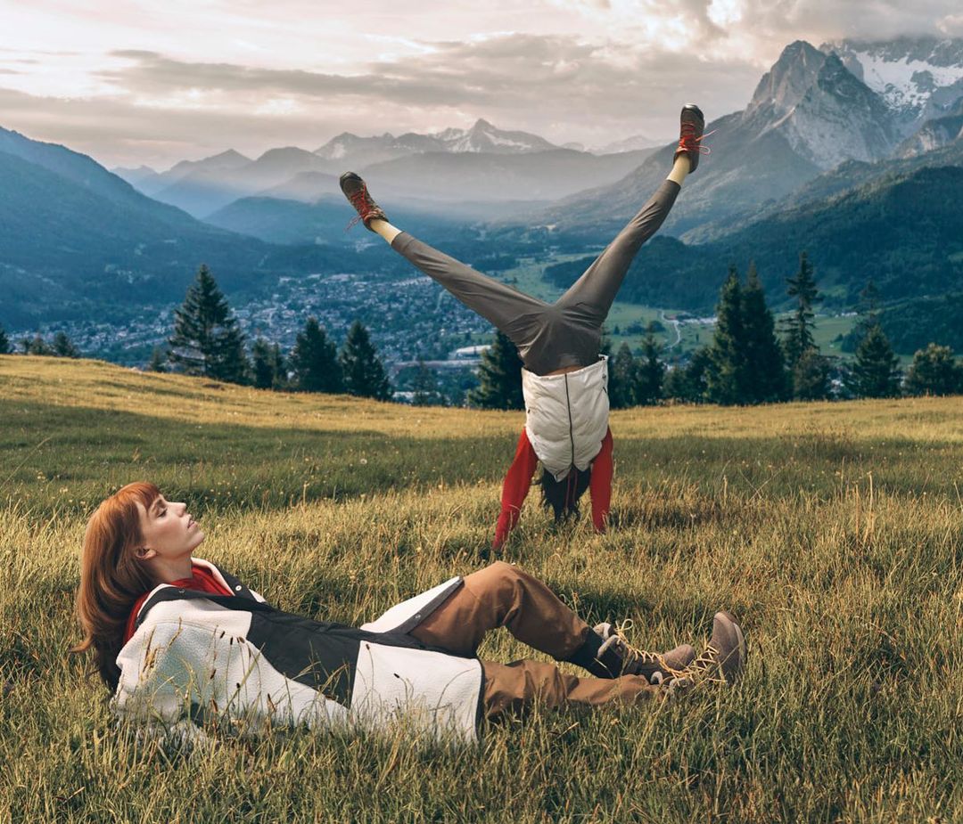 Two women in a grassy field with mountains in the background. One woman is lying down, while the other is doing a handstand.
