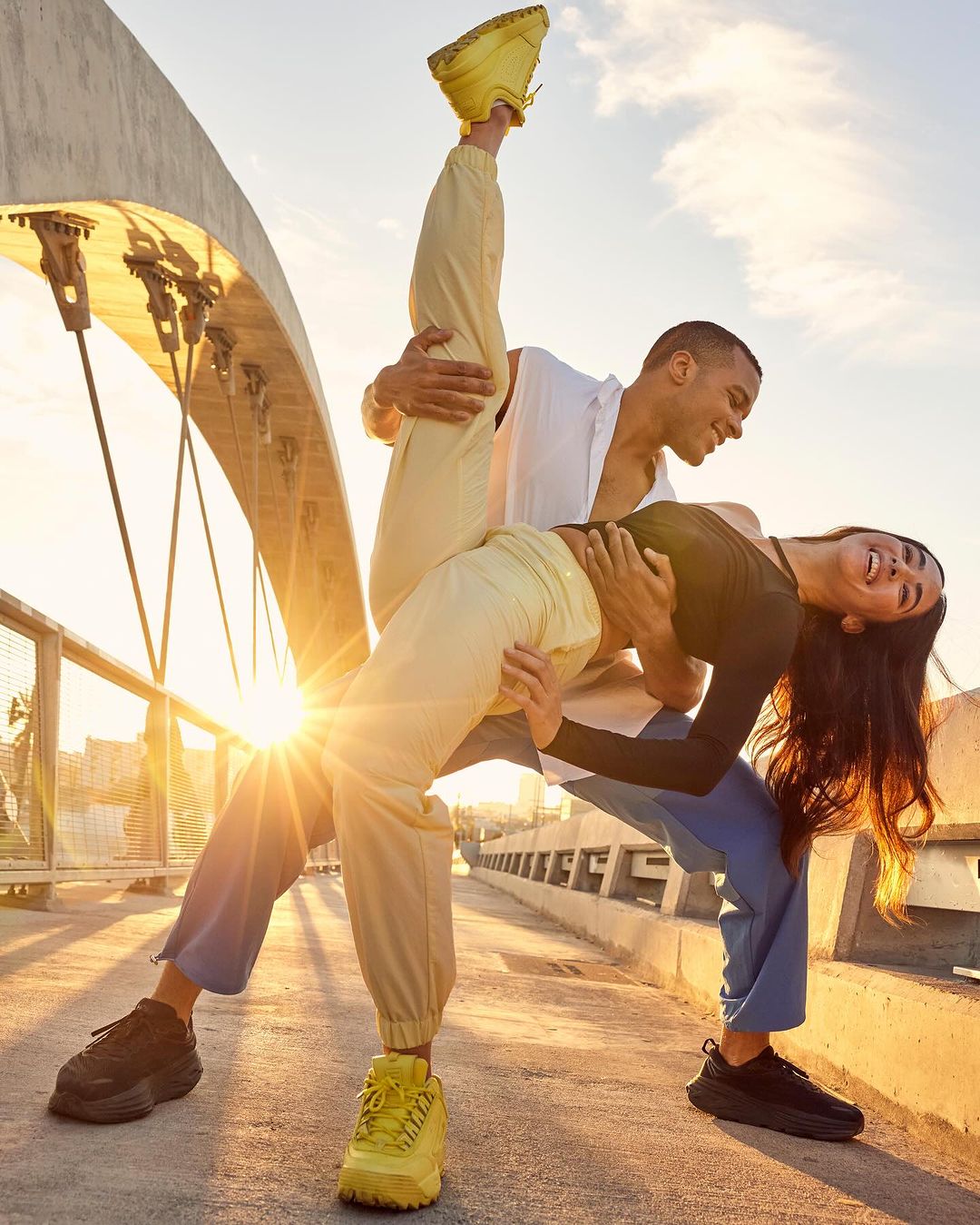 Two athletic adults in sportswear pose on a bridge at sunset with a bright sun flare. The couple is dancing in sneakers.