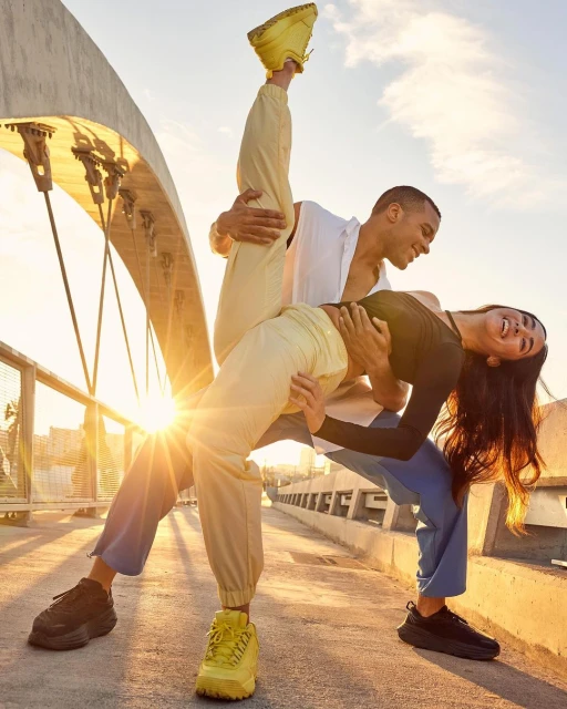 Two athletic adults in sportswear pose on a bridge at sunset with a bright sun flare. The couple is dancing in sneakers.
