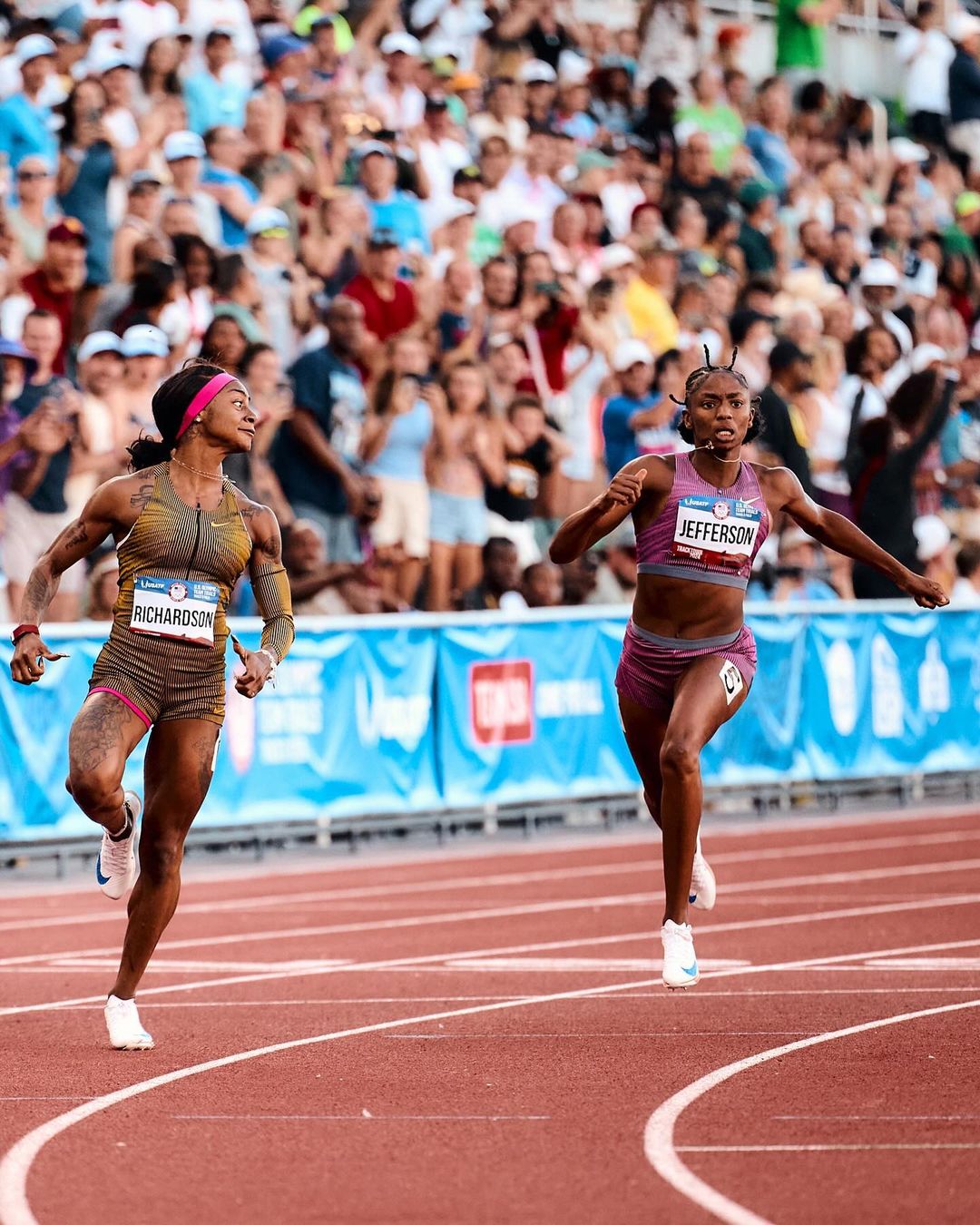 Two adult female athletes, Richardson and Jefferson, sprint on an outdoor track during a race, cheered on by a large crowd of spectators.