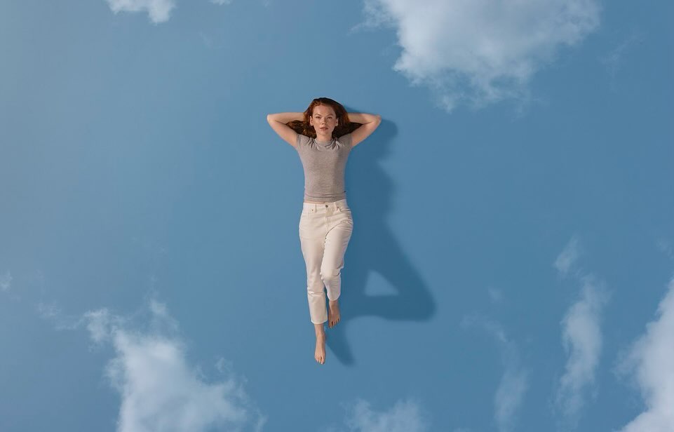 Young woman lying on her back in a sky full of clouds. She is wearing a gray top and white pants with hands behind her head.