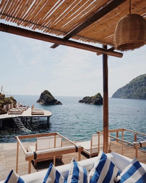 Scenic coastal view from a seaside lounge featuring lounge chairs on a deck, with islands in the distance, and a bamboo-covered pergola.