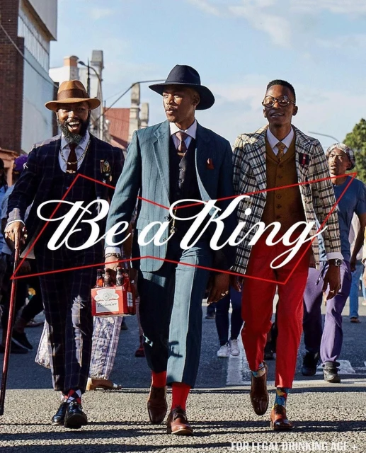 Three well-dressed men walk on an urban street holding a six-pack of beer, wearing hats, suits, and ties, with blurred people in background.