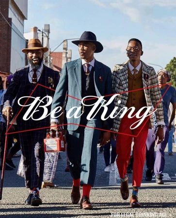 Three well-dressed men walk on an urban street holding a six-pack of beer, wearing hats, suits, and ties, with blurred people in background.
